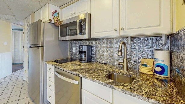 A kitchen with white cabinets, a stainless steel refrigerator, a microwave, a coffee maker, and a sink with a gold faucet. - St Clement's Unit 409 (Myrtle Beach)