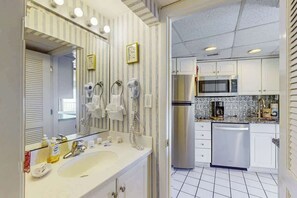 A bathroom with a sink, mirror, and towel racks. Adjacent is a kitchen with a microwave, refrigerator, and dishwasher. - St Clement's Unit 409 (Myrtle Beach)