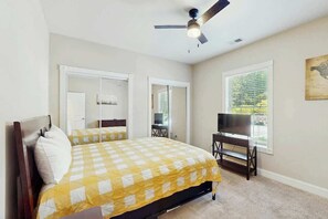 Ein Schlafzimmer mit einem Bett, Deckenventilator, einem Fernseher auf einem Schrank und einem Fenster mit Jalousien.