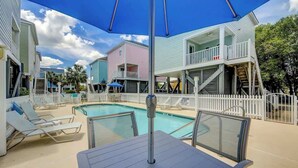 A pool area with lounge chairs and umbrellas, surrounded by a white fence and colorful buildings. - The Tickled Pink Retreat (Murrells Inlet)