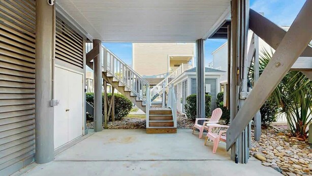 A covered outdoor area with a concrete floor, a staircase leading to a second level, and a pink chair. - The Tickled Pink Retreat (Murrells Inlet)