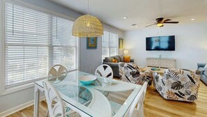 A modern living room with a glass dining table, white chairs, a patterned sofa, a ceiling fan, and a flat-screen TV. - The Tickled Pink Retreat (Murrells Inlet)