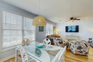 Ein modernes Wohnzimmer mit einem Glastisch, weißen Stühlen, einem gemusterten Sofa, einem Deckenventilator und einem Flachbildfernseher.
