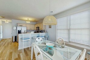 A modern kitchen with white cabinetry, a dining area with a glass table, and a large window with blinds. - The Tickled Pink Retreat (Murrells Inlet)