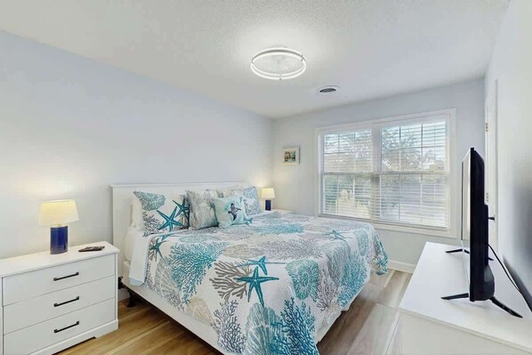 A bedroom with a bed, bedside table, lamp, and a TV. - The Tickled Pink Retreat (Murrells Inlet)