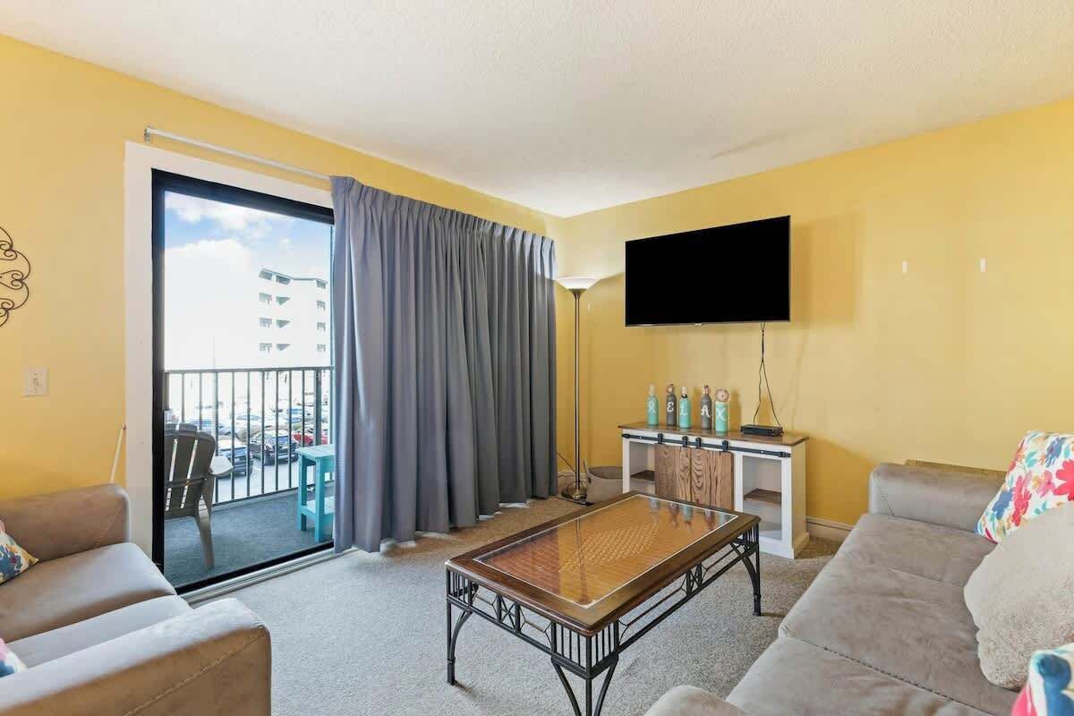 Ett vardagsrum med en soffa, ett soffbord, en platt-TV, en balkong med utsikt över bilar och byggnader samt ett litet bord med flaskor.