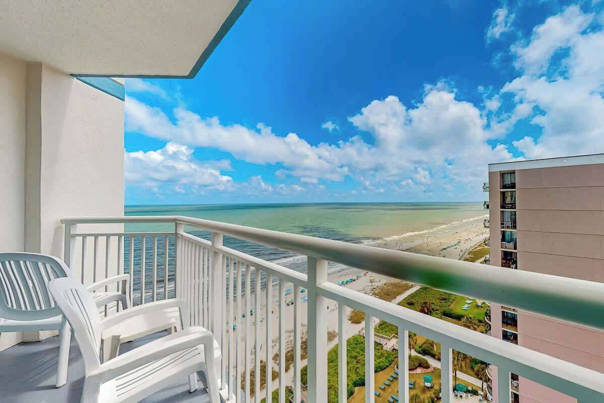 Un balcon donnant sur une plage et l’océan, des meubles d’extérieur blancs et un immeuble à plusieurs étages.