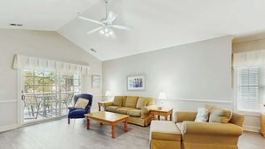 A spacious living room with a high ceiling, a ceiling fan, a sofa set, a coffee table, and a dining area with a table and chairs. - Magnolia Pointe Unit 305 (Myrtle Beach)
