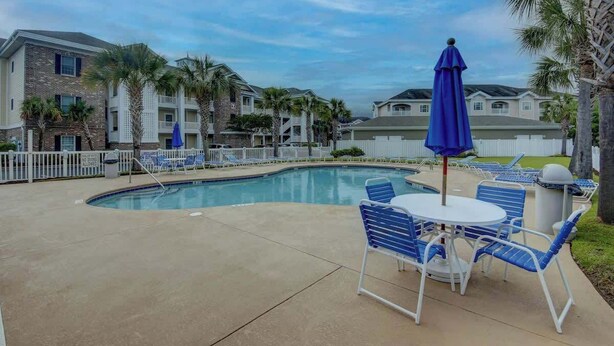 A swimming pool with lounge chairs and umbrellas, surrounded by a fence, with buildings in the background. - Magnolia Pointe Unit 305 (Myrtle Beach)