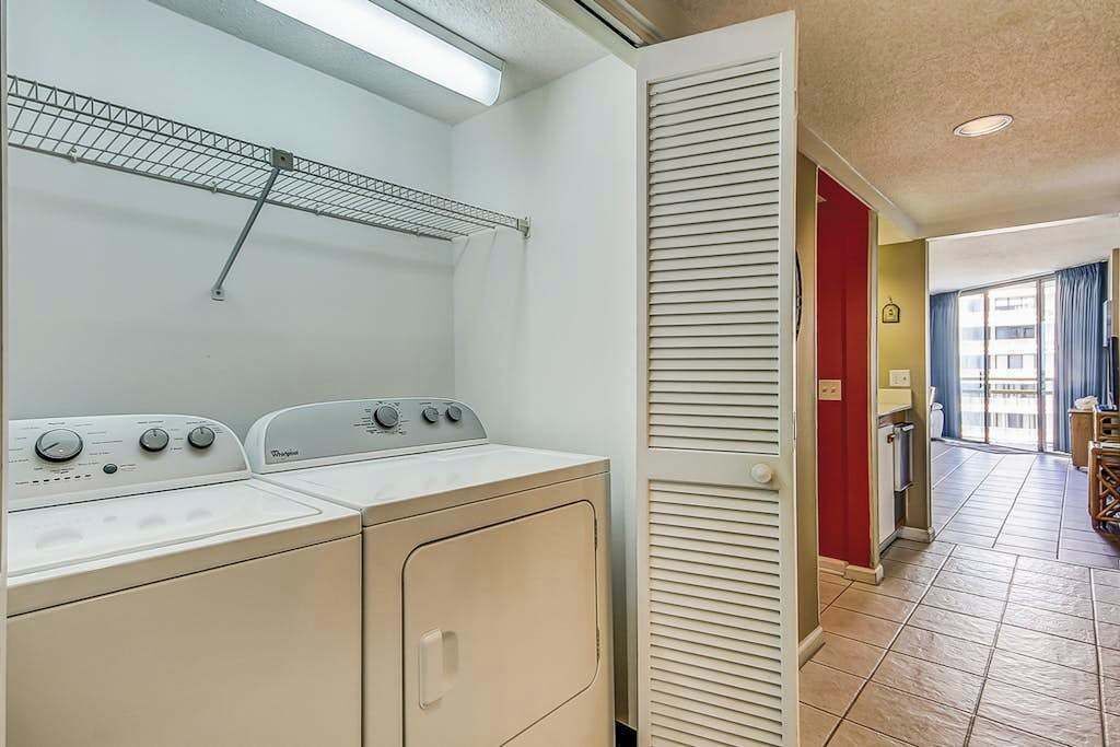 Een wasruimte met een wasmachine en droger, een metalen rek en een deur naar een kamer met een rode wand en een raam.