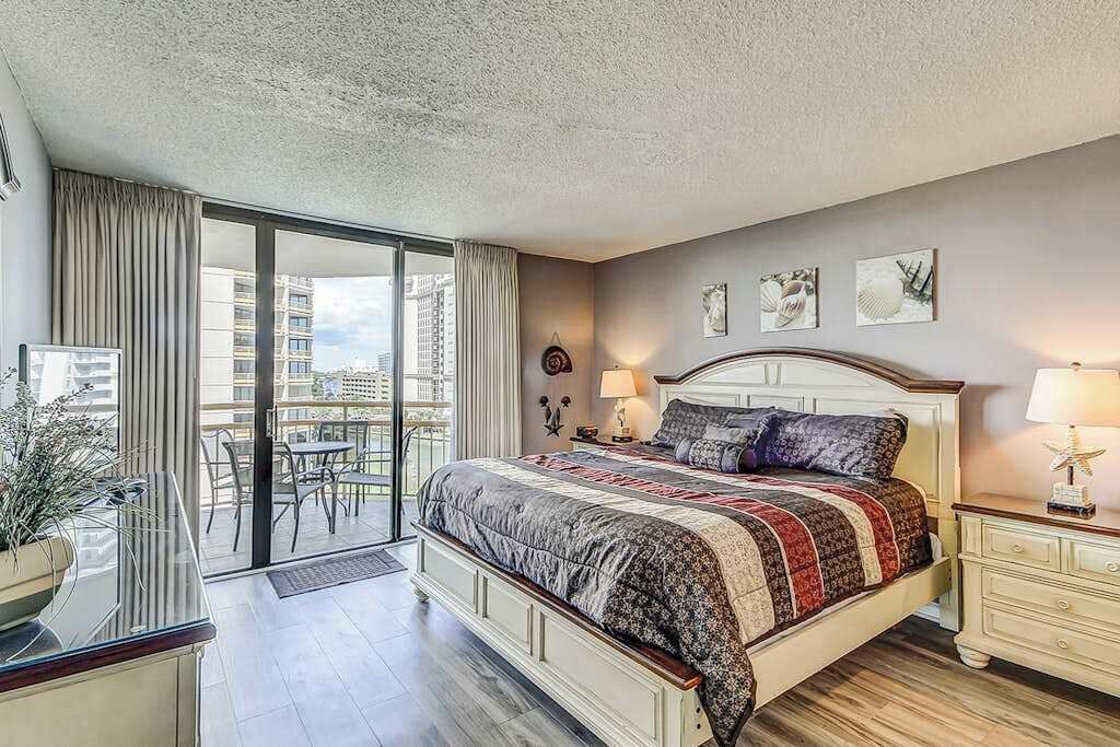 A bedroom with a large bed, a nightstand, a lamp, a potted plant, and a view of the city through a sliding door.