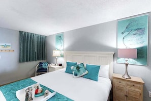 A hotel room with a bed, a nightstand, a lamp, a chair, and artwork on the walls. - Bay Watch Resort Unit 0334 (North Myrtle Beach)
