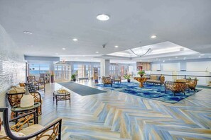 A spacious lobby with a herringbone-patterned floor, seating areas with wicker furniture, and a view of the sea through large windows. - Bay Watch Resort Unit 0334 (North Myrtle Beach)