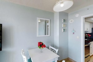 A room with a white table, a chair, a vase with flowers, a mirror, and a door leading to another room. - Bay Watch Resort Unit 0334 (North Myrtle Beach)