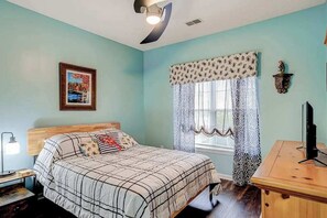 Ein Schlafzimmer mit einem Bett, einem Nachttisch, einer Lampe, einem Fenster mit gemusterten Vorhängen und einem an der Wand montierten Fernseher.