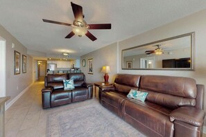 A living room with a brown leather sofa, a ceiling fan, a large mirror, and a kitchen in the background. - Bahama Sands Unit 0706 (North Myrtle Beach)