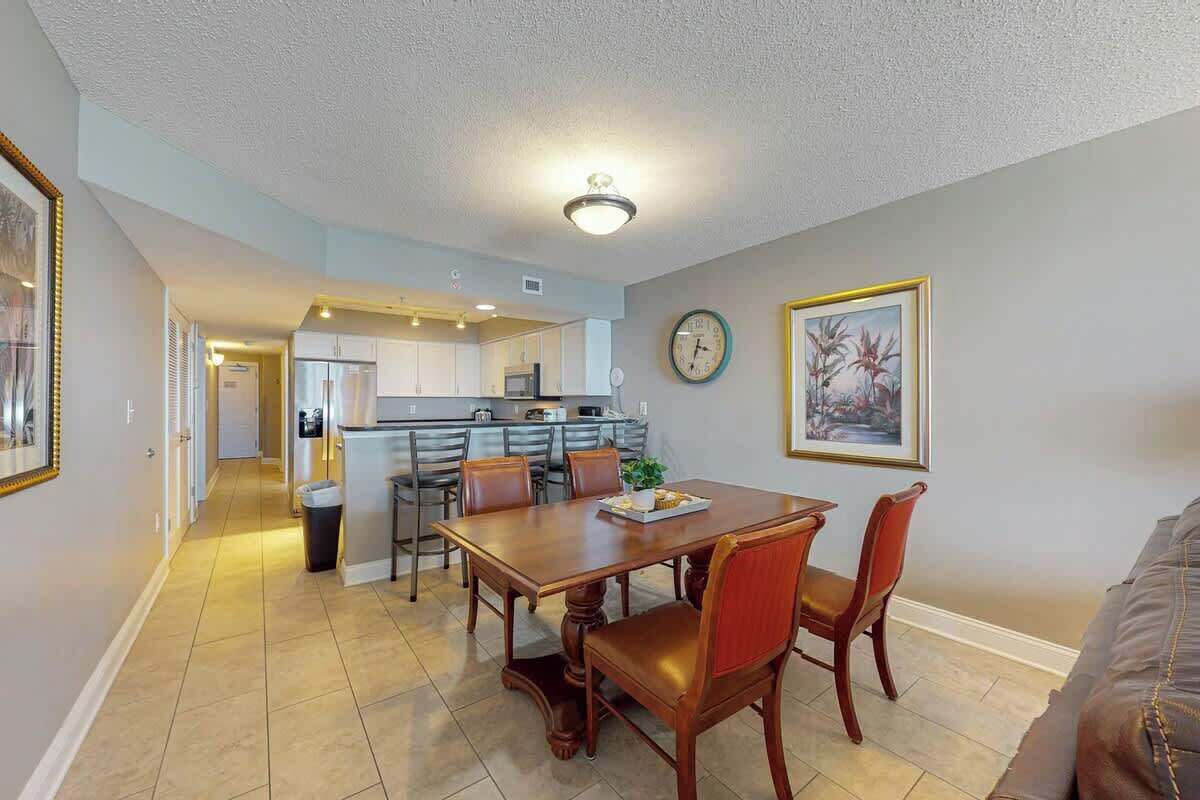 A dining area with a wooden table and chairs, a kitchen in the background, and a clock on the wall.