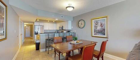 A dining area with a wooden table and chairs, a kitchen in the background, and a clock on the wall.