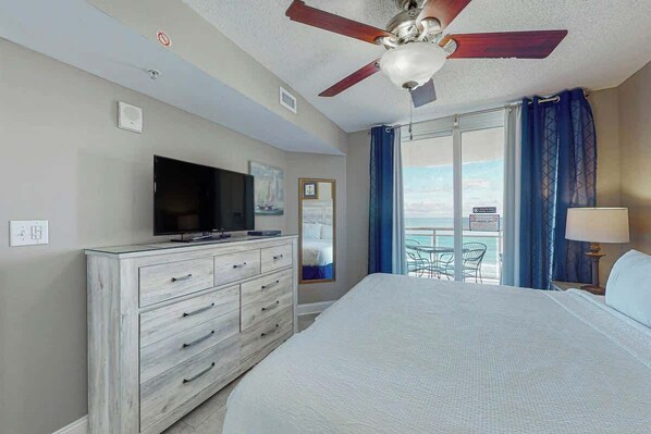 Quarto de hotel com cama, televisão em uma cômoda, ventoinha no teto e vista para o exterior.