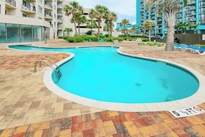 A swimming pool with a tiled deck, surrounded by palm trees and a multi-story building. - Palace Resort Unit 1210 (Myrtle Beach)
