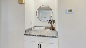 A bathroom with a white vanity, a round mirror, a small potted plant, and a thermostat on the wall. - Pelicans Watch Unit 121 (Myrtle Beach)