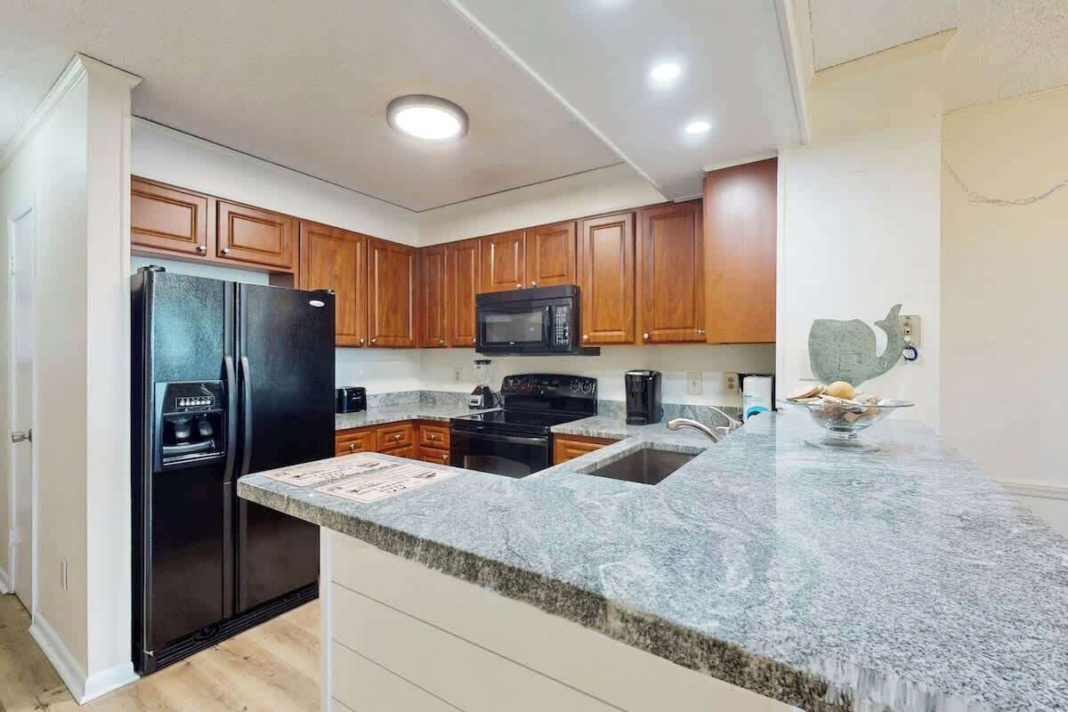 A kitchen with wooden cabinets, a black refrigerator, a microwave, a sink, and a granite countertop.