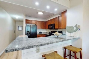 A kitchen with granite countertops, wooden cabinets, a refrigerator, a microwave, and two bar stools. - Pelicans Watch Unit 121 (Myrtle Beach)
