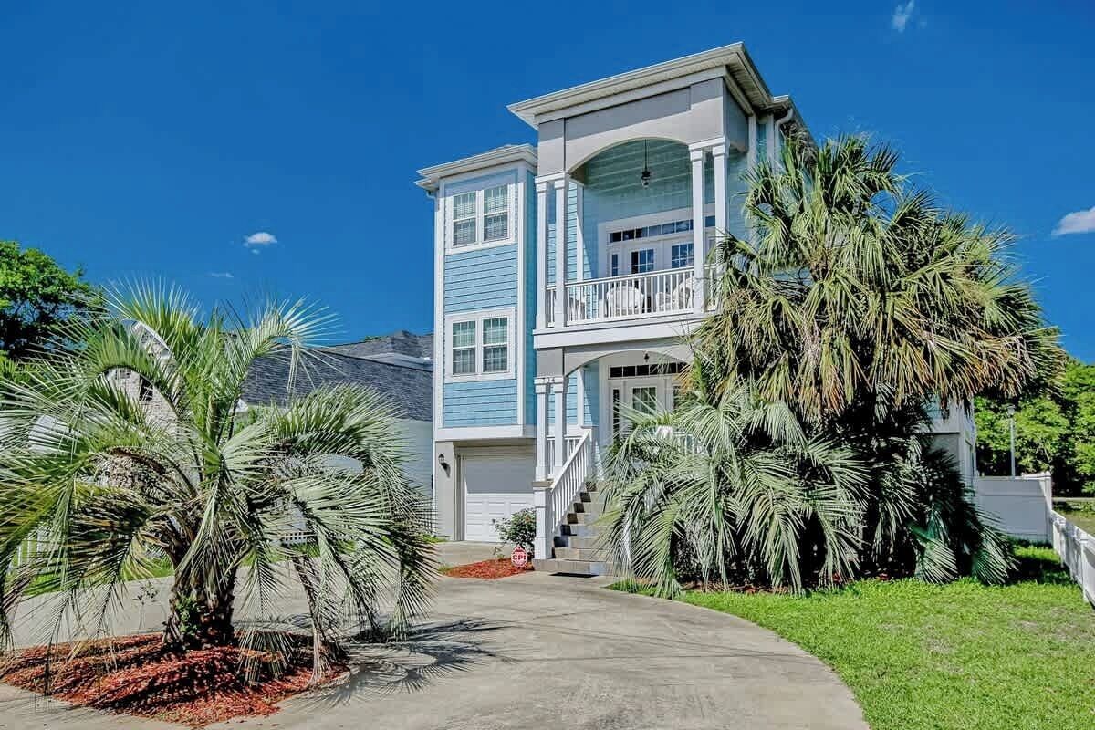 A two-story house with a balcony, surrounded by palm trees and a clear sky.