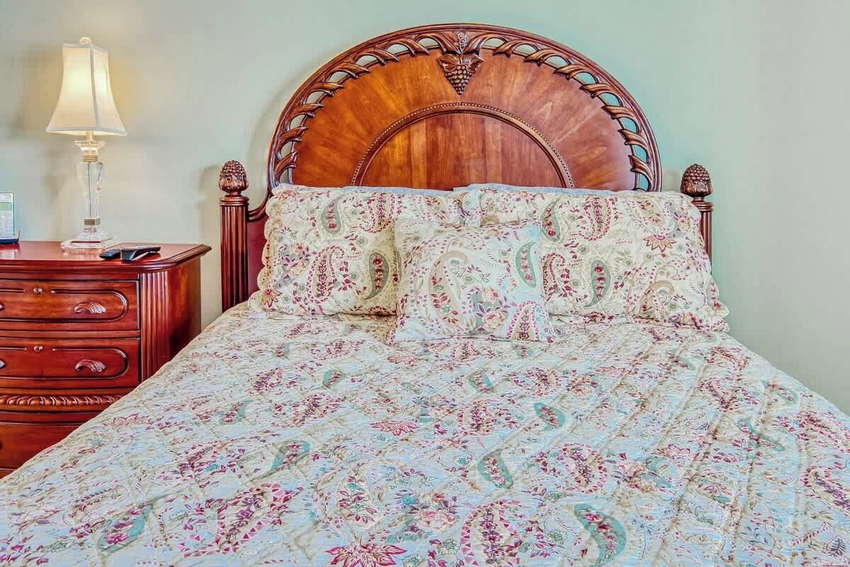 A bed with a floral patterned bedspread and matching pillows, a wooden headboard, and a bedside table with a lamp.