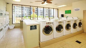A laundry room with multiple washing machines, a closed sign, and a view of a residential building through large windows. - Myrtle Beach Resort B-349 (Myrtle Beach)