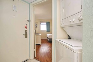 A laundry room with stacked washers and dryers, a door with a fire alarm, and a hallway leading to a bedroom with a window and curtains. - Bay Watch Resort 1019 (North Myrtle Beach)