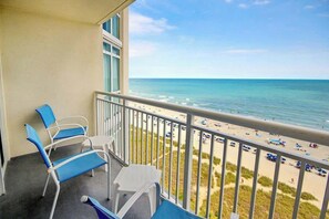 A balcony with a view of the beach, featuring a railing, a table, and two chairs. - Bay Watch Resort 1019 (North Myrtle Beach)