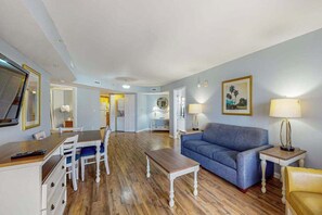 A living room with a blue sofa, a wooden coffee table, and a dining area with white chairs. - Ocean Reef Resort 723 (Myrtle Beach)