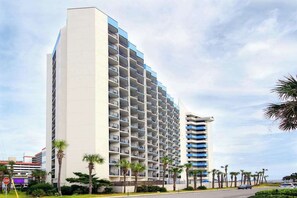 Ein mehrstöckiges Hotel mit modernem Design, mit Balkonen auf jeder Etage, in der Nähe einer von Palmen gesäumten Straße.