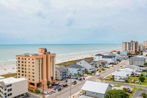 A coastal town with a beachfront hotel, a road with parked cars, and buildings in the background. - Wellington Unit 304 Condo (North Myrtle Beach)