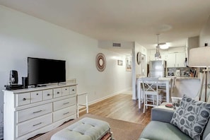 A living room with a flat-screen TV, a white dresser, a grey sofa, and a kitchen area with white cabinets and a dining table. - Salty Mermaid at Wind Crest Villas (North Myrtle Beach)