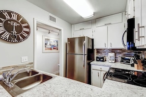 A modern kitchen with stainless steel appliances, a double sink, and a large wall clock. - Salty Mermaid at Wind Crest Villas (North Myrtle Beach)