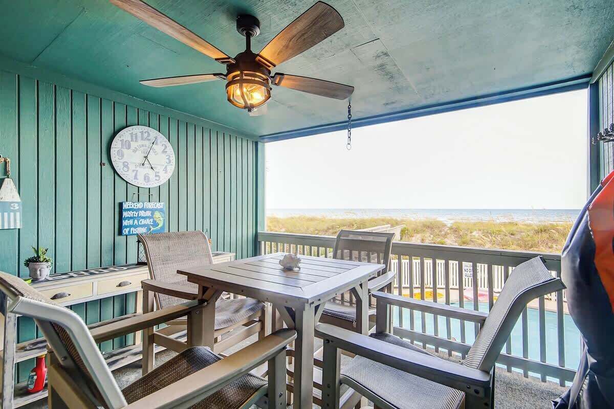 A covered outdoor seating area with a table and chairs, a ceiling fan, a clock, and a view of the beach.