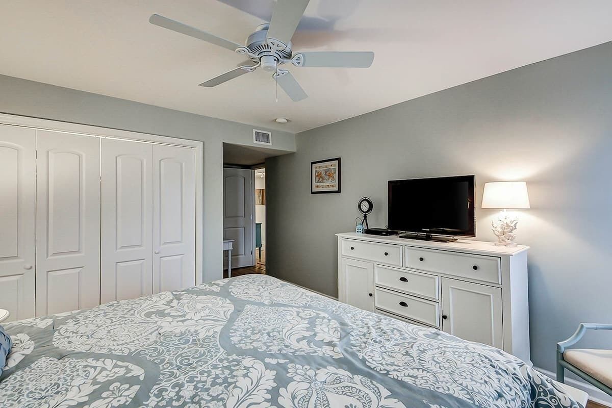 A bedroom with a bed, a television, a dresser, a ceiling fan, and a door leading to another room.