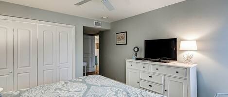 A bedroom with a bed, a television, a dresser, a ceiling fan, and a door leading to another room.