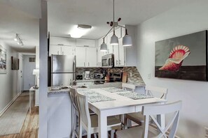 A modern kitchen with white cabinetry, a dining area with a white table and chairs, and a hanging light fixture. - Salty Mermaid at Wind Crest Villas (North Myrtle Beach)