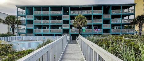 A multi-story building with balconies, a wooden walkway leading to the entrance, and a palm tree in the foreground.