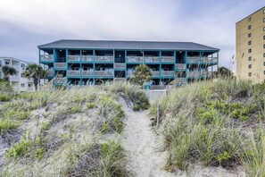 A beachfront building with multiple balconies, a sandy path leading to it, and surrounding vegetation. - Salty Mermaid at Wind Crest Villas (North Myrtle Beach)