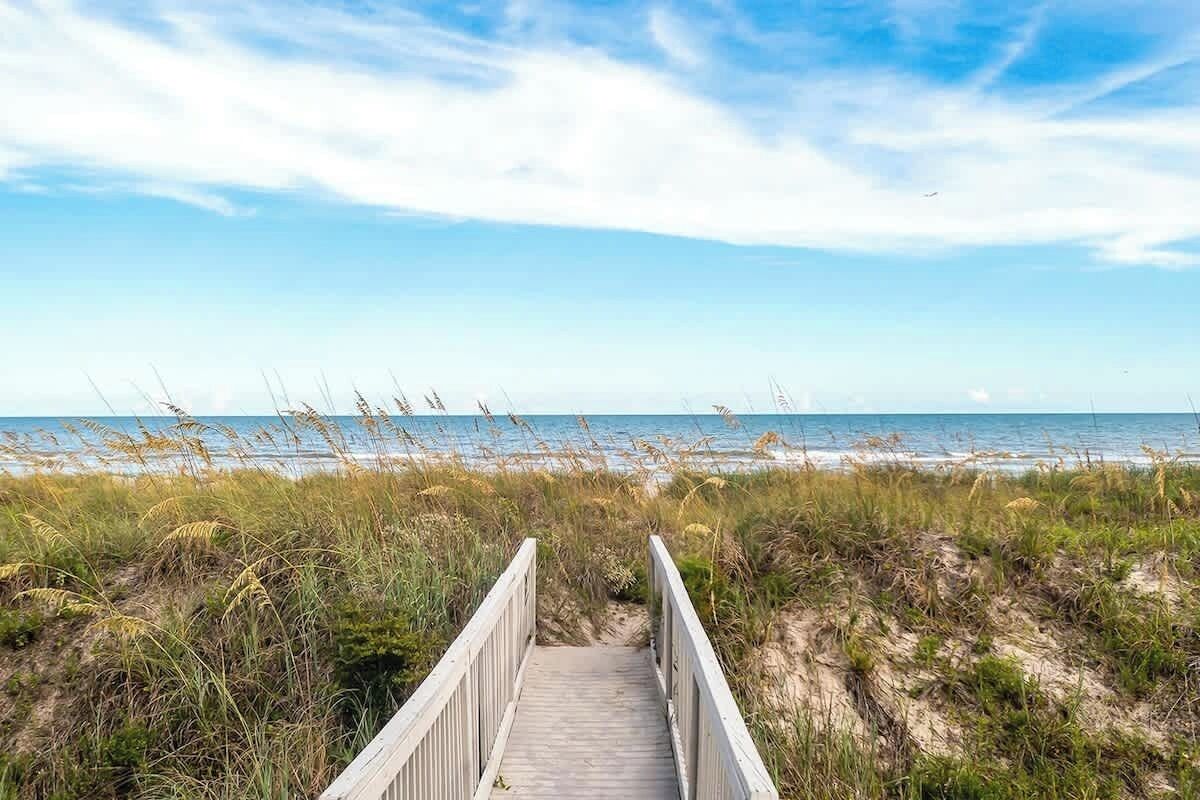 A wooden boardwalk leading to a beach with dunes and grasses.