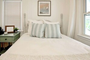 A neatly made bed with striped pillows, a framed picture above it, a bedside table with a plant, and a window with blinds. - Lovely Beach Bungalow (North Myrtle Beach)