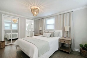 A bedroom with a large bed, bedside tables, a chandelier, and a view of a bathroom. - Lovely Beach Bungalow (North Myrtle Beach)
