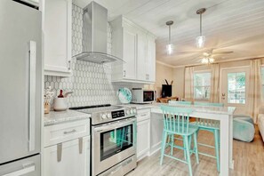 A kitchen with white cabinetry, a stainless steel range, and a microwave. There are turquoise bar stools and a window with curtains. - Lovely Beach Bungalow (North Myrtle Beach)