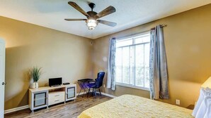 Quarto de hotel com cama, escrivaninha com cadeira, ventilador de teto e janela com cortinas.