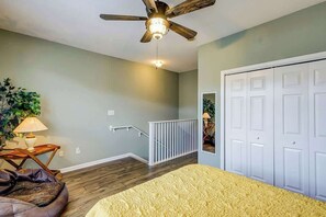 Ein Schlafzimmer mit Deckenventilator, einem Bett mit gelber Bettdecke, einem Nachttisch mit Lampe, einer Pflanze und einer Treppe zu einem Schrank.