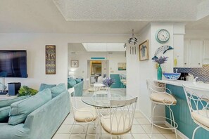 Ein modernes Wohnzimmer mit einer Couch, einem Glastisch und Stühlen. Es gibt eine Küchenzeile mit einer Arbeitsplatte und einer Uhr an der Wand.